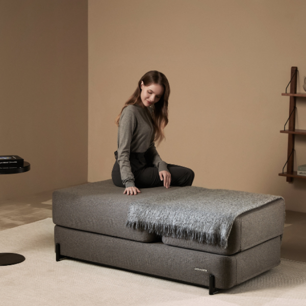 Woman sitting on a gray Ceragem S4 massage bed in a room with a shelf and small table.