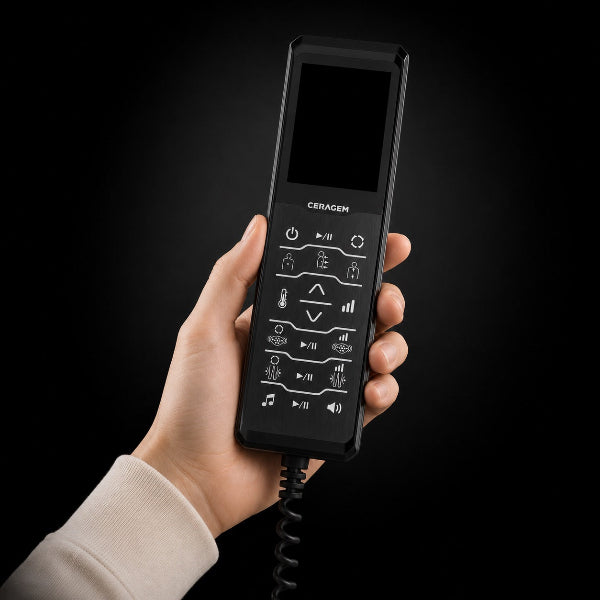Hand holding a black Ceragem wired remote with a digital screen and illuminated control buttons against a dark background, with a coiled cord extending from the bottom.
