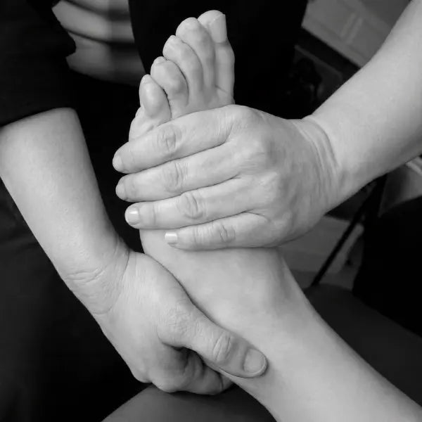 Black-and-white close-up of hands performing a foot massage, focusing on the ankle and sole.
